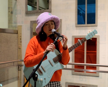 A person wearing a mauve hat and orange top, holding a light blue guitar, sings into a microwave.
