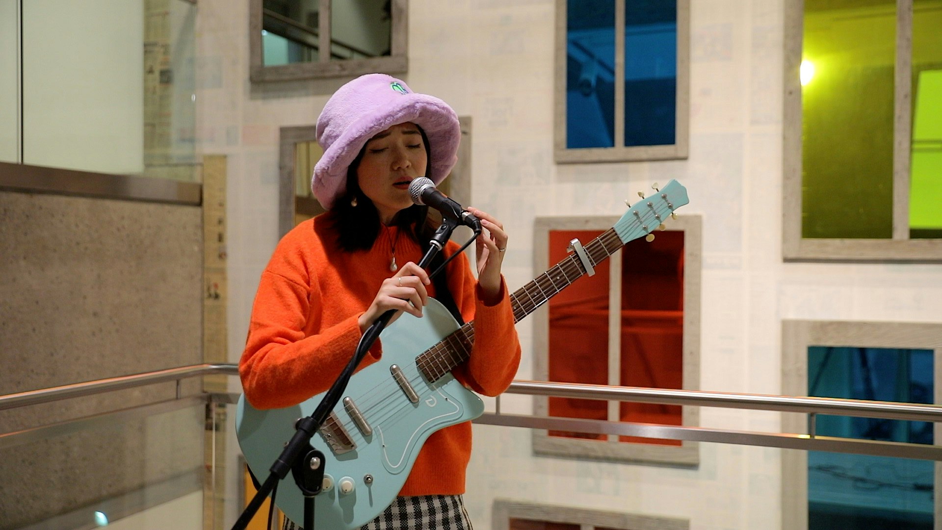 A person wearing a mauve hat and orange top, holding a light blue guitar, sings into a microwave.
