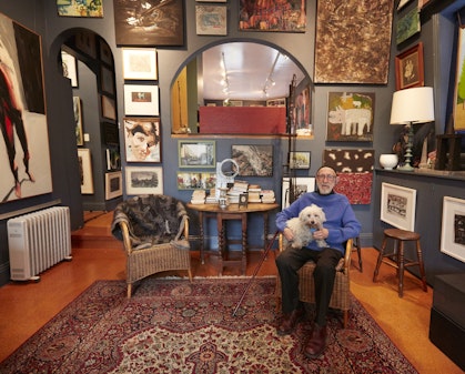 A man sits with a small dog in his lap in a room in which the walls are densely hung with artworks.