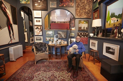A man sits with a small dog in his lap in a room in which the walls are densely hung with artworks.
