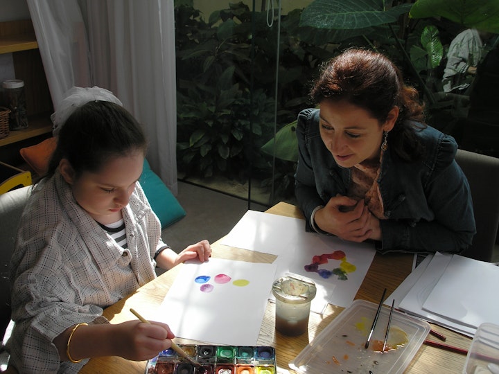 A young girl sits at a table painting a sheet of paper while an adult looks on.