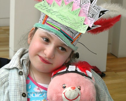 A girl wearing a colourful paper and feather headress hugs a pink stuffed toy which is wearing a similar headdress.