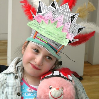 A girl wearing a colourful paper and feather headress hugs a pink stuffed toy which is wearing a similar headdress.