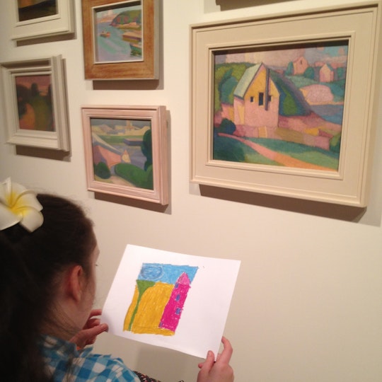 A girl studies an illustration in front of several colourful landscapes hanging on a wall.