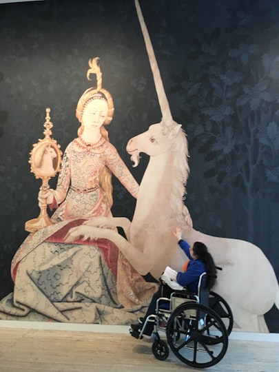 A person in a wheelchair reaches out a hand to a large unicorn on a wall. With the unicorn is a seated lady.