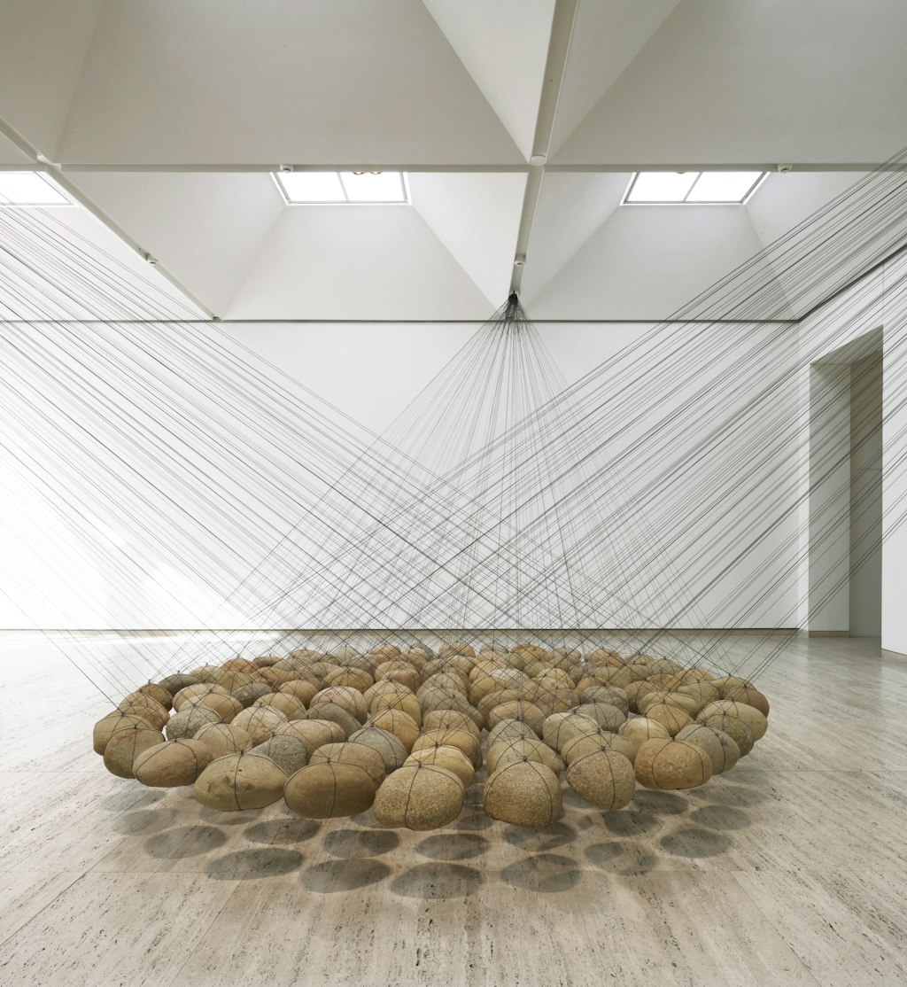 Dozens of smooth rounded stones are suspended on wires from the ceiling so that they hang in cluster just above a gallery floor.
