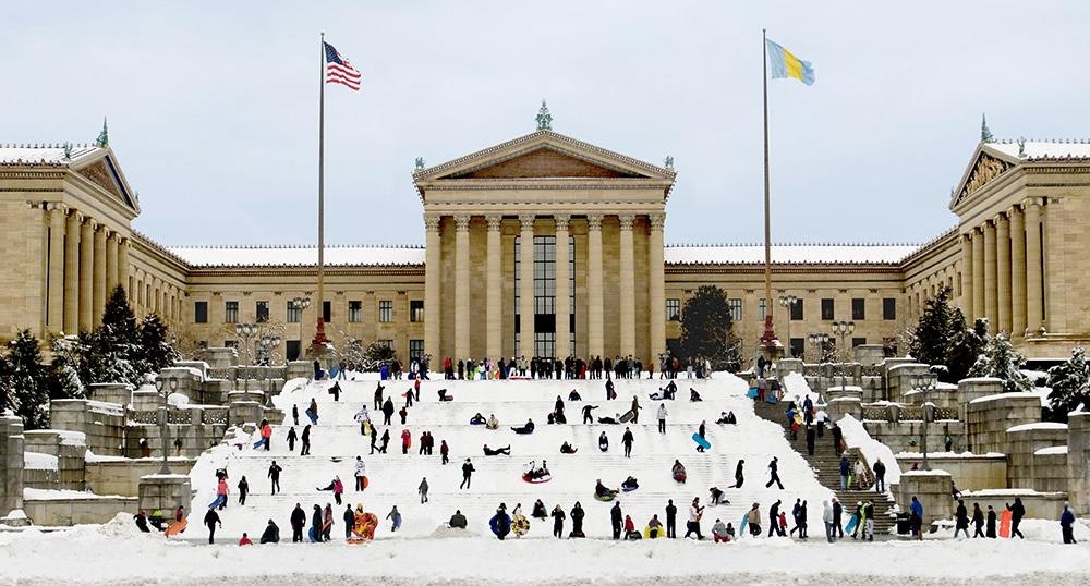A large official-looking building with collonaded porticos at the top of a long flight of snow-covered steps, dotted with people.