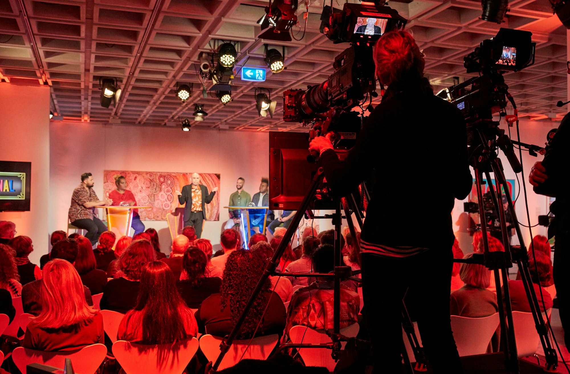 Five people on stage watched by a seated audience and filmed by a TV cameraperson.