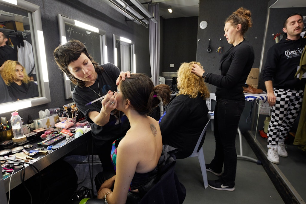 Two people sit in front of mirrors and a table laden with make-up while having make-up applied and hair done by two standing people, with another person standing to the side.