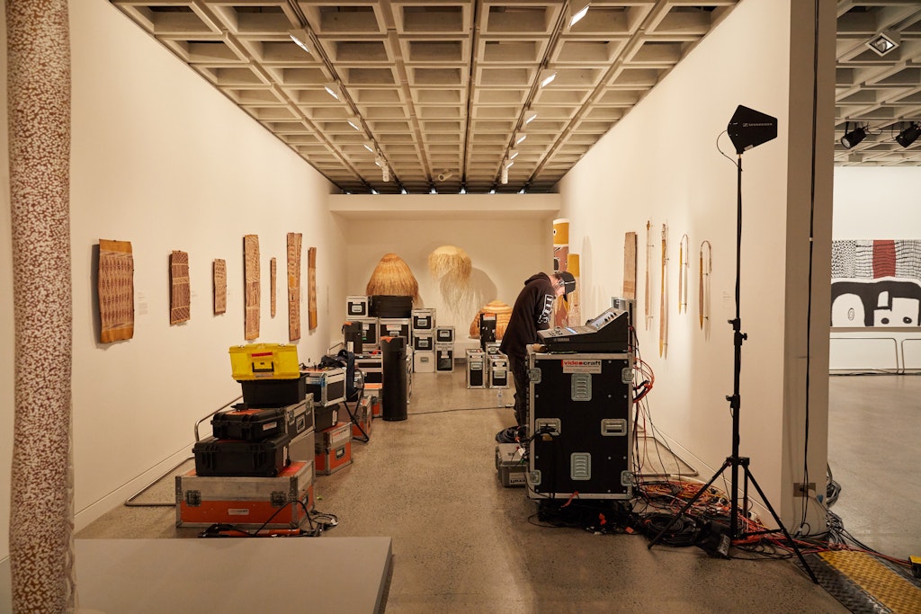 In a gallery space hung with Aboriginal art, there are stacks of TV equipment and a person leaning over one of the pieces.