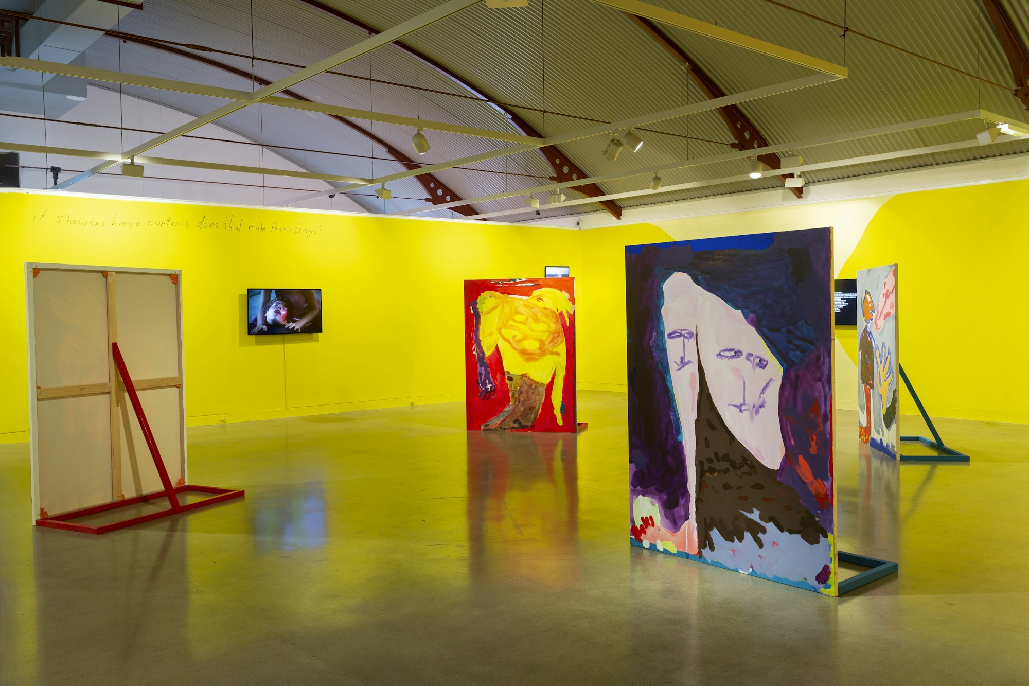 Four large paintings are propped up on a gallery floor. Around them are partial-height  walls painted bright yellow on which hang a few video screens.