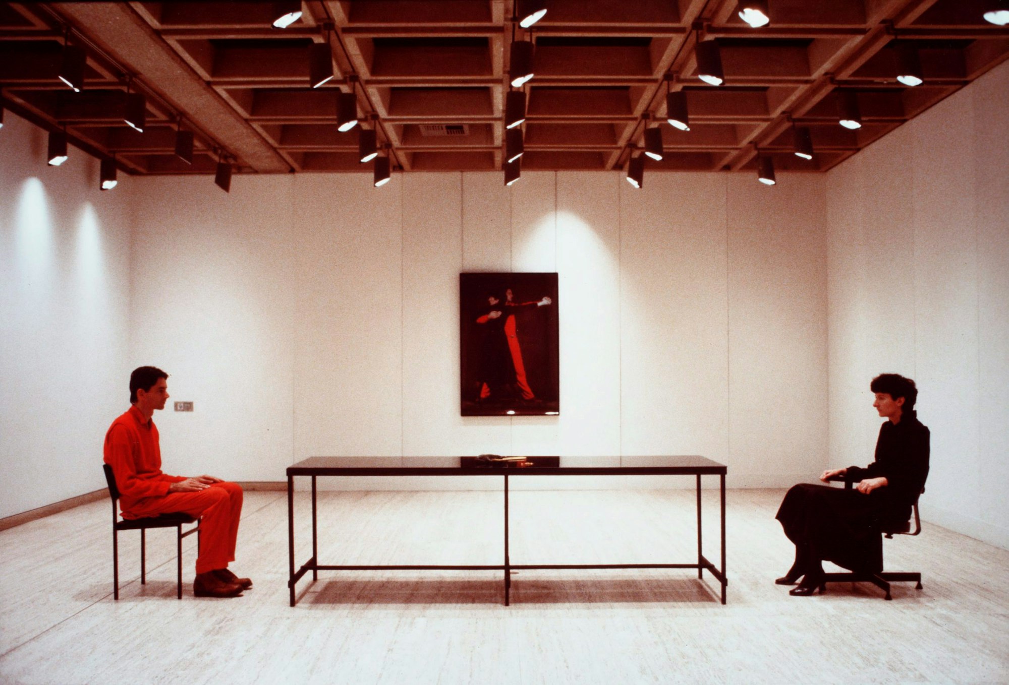In a gallery space, two people sit looking at each other from opposite ends of a long table. An artwork hangs on the far wall.