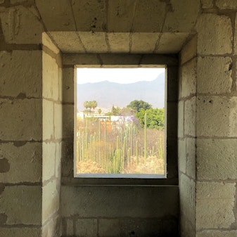 A window in a room constructed from large cement bricks looks out over a field of cacti to distant buildings, trees and hills.