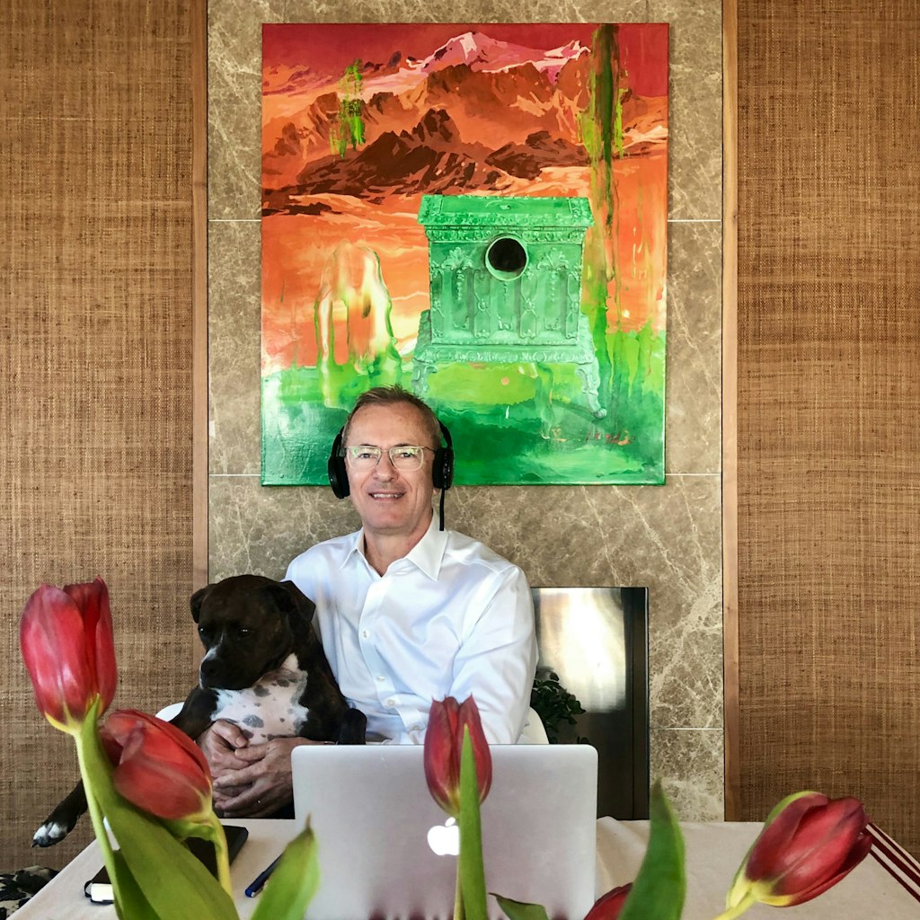 A person wearing headphones sits with a dog in front of a laptop computer on a table. In the foreground are some flowers. In the background is an otherworldly landscape painting.