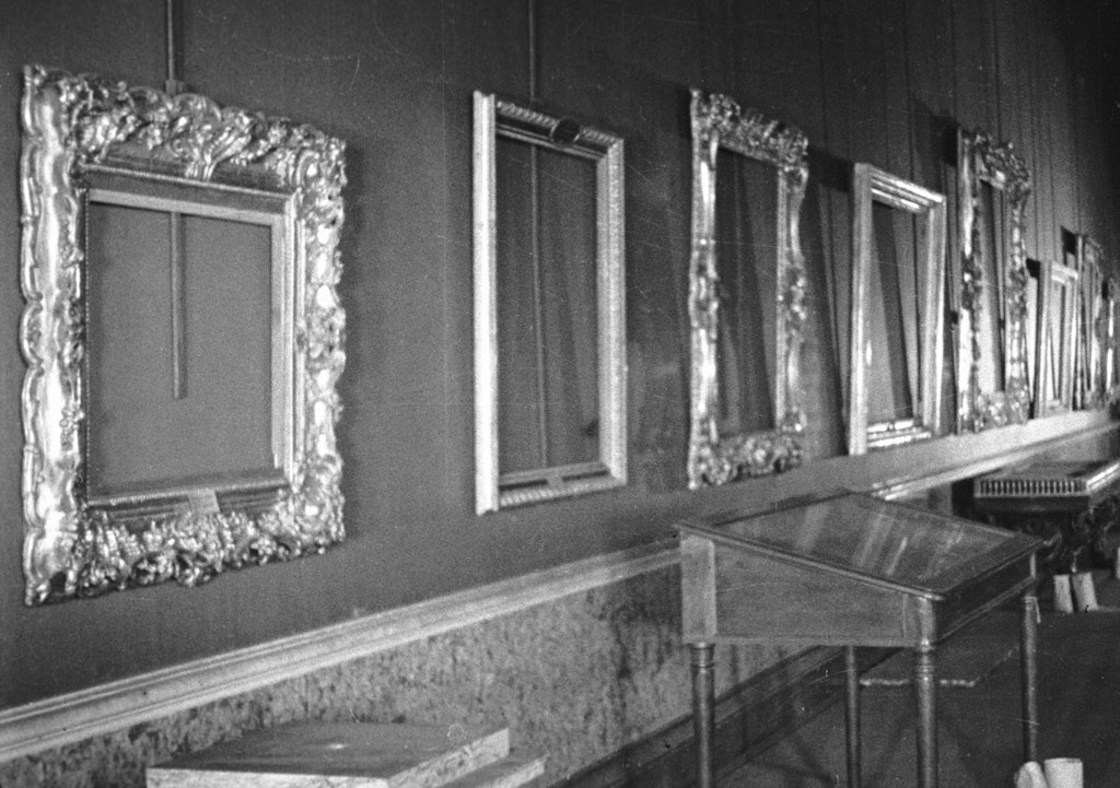 A black-and-white photo of a line of empty ornate frames along a wall with small display cases in front of them.