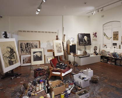 A cluttered room, with an armchair in the centre, surrounded by painting materials on the floor and in boxes. Around the edges of the room are artworks and a bookshelf.
