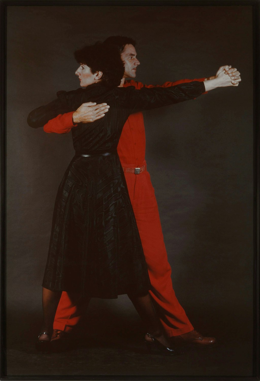 Two people, one in red pants and shirt, the other in a black skirt and shirt, in a tango dance pose.
