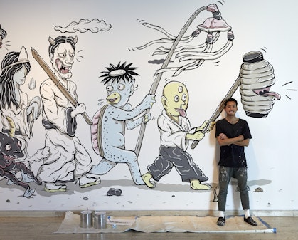 A person stands on a drop sheet with two paint tins in front of a mural of supernatural Japanese characters.