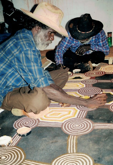 Two Aboriginal men sitting cross-legged on the floor on top of a large artwork that they are painting with small brushes.