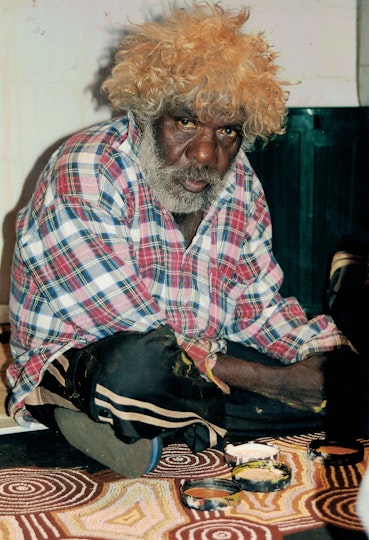 An Aboriginal men sitting cross-legged on the floor on top of a large painting, with jar lids of paint in front of him.