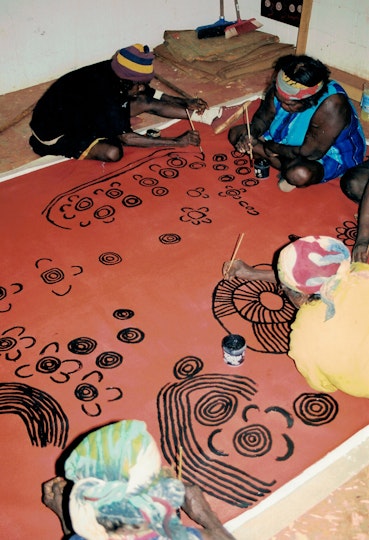 Four Aboriginal women on the floor on top of a large artwork that they are painting with small brushes.