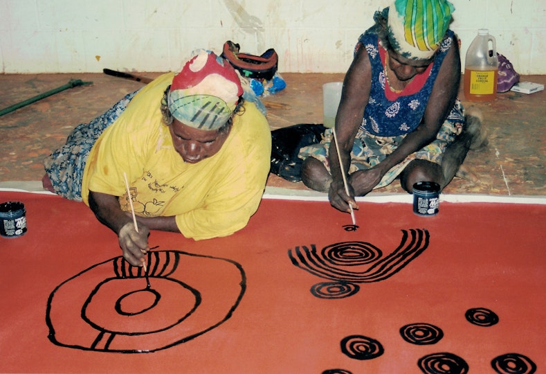 Two Aboriginal women on the floor on top of a large artwork that they are painting with small brushes.