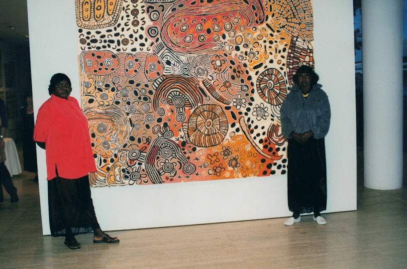 Two Aboriginal women stand with a large painting on a gallery wall.