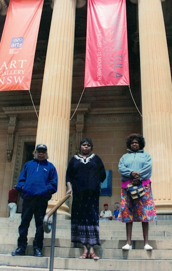 Three Aboriginal people stand on steps in front of tall columns, between which hang two banners. One banner has a logo consisting of the five Olympic rings with 'arts' written underneath, and a logo consisting of 'Art Gallery NSW'; the other says 'Papunya Tula Genius & Genesis 18 August - 12 November'..