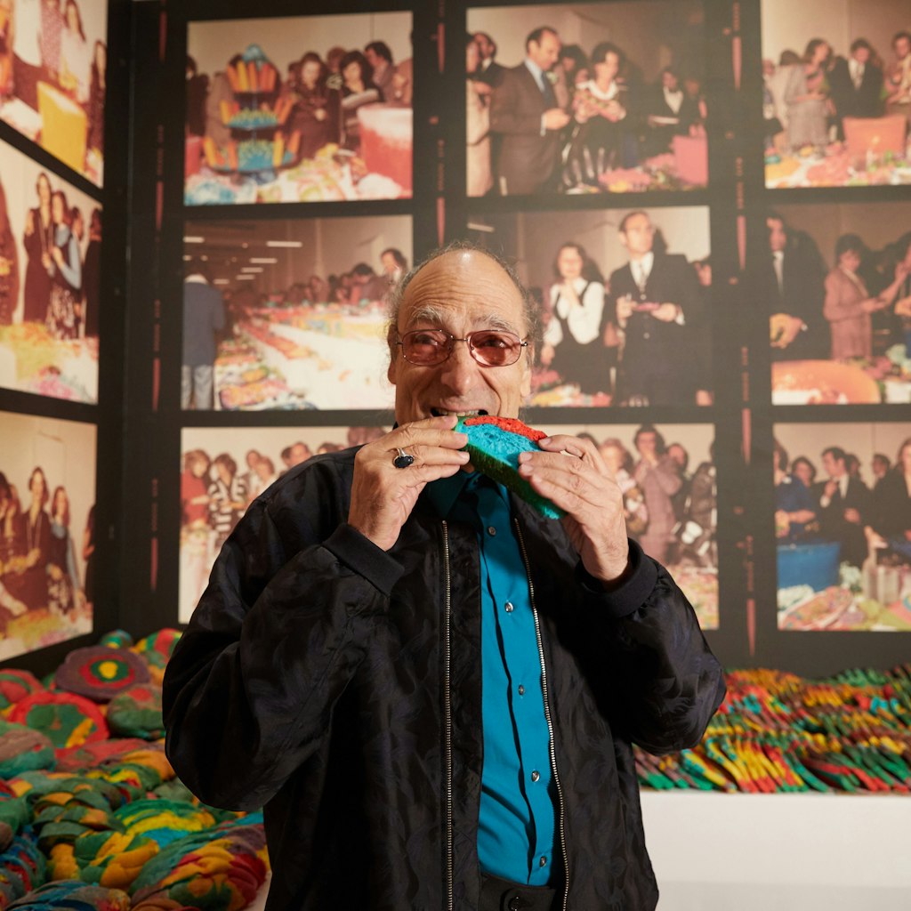 A person bites into a piece of bright blue and red bread among a gallery display of many pieces of multi-coloured bread on plinths and photos on the walls.