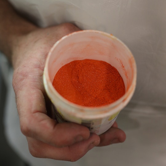 A hand holds a white plastic pot of orange powder.