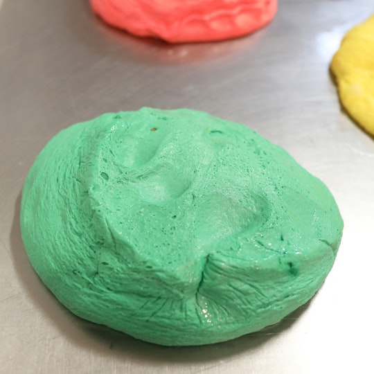 A sphere of green dough sits on a silver-coloured metal sheet, with yellow and orange lumps of dough in the background.