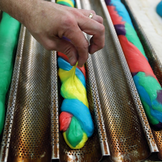 A hand adjusts a multi-coloured length of dough in a grooved, perforated metal tray.