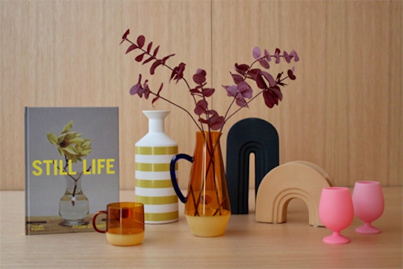 Book titled 'Still Life', orange glass mug, lime green and white striped vase, orange glass jug with black handle and dried leaves arranged in it, black book end and bright pink silicone wine globlets.