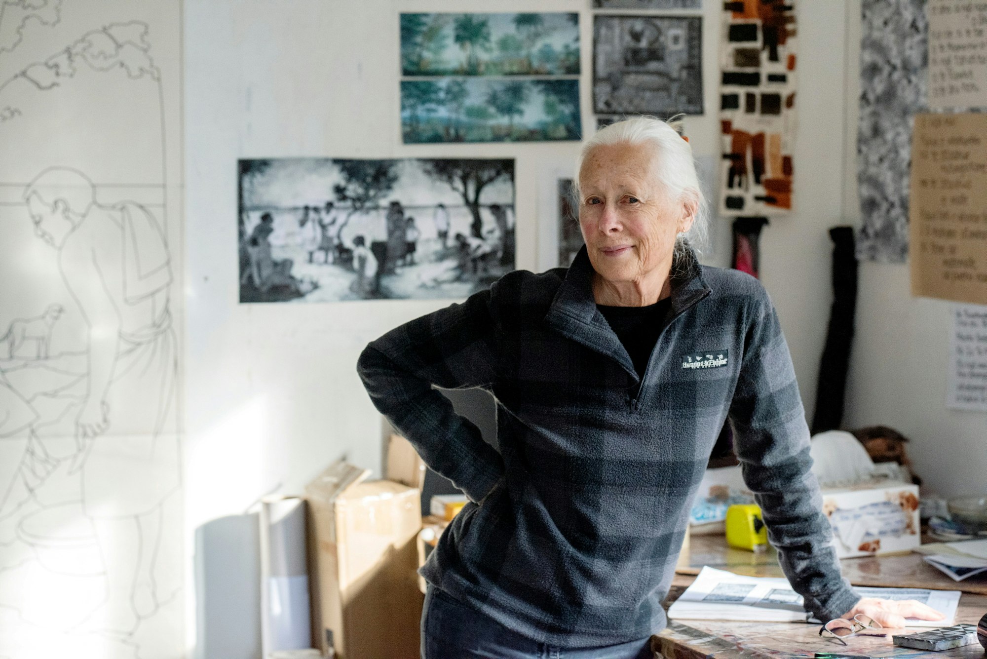 A person with hair pulled back from their face, wearing jeans and a long-sleeved top, leans on an object-strewn table with one hand, the other is on their hip. Art, images and written sheets of paper hang on the walls behind her.