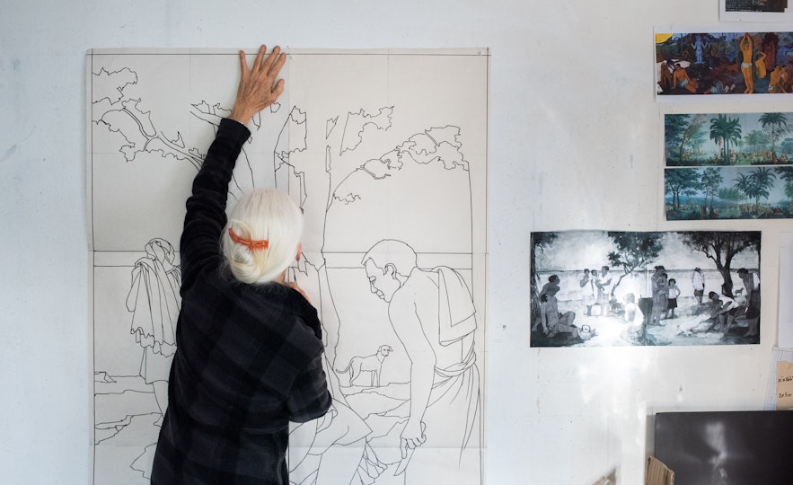A person reaches up to the top of a large drawing on a wall. Other images are fixed to the wall on its right.