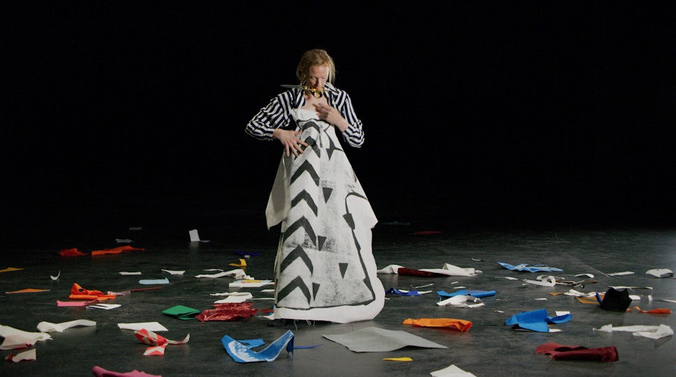 A person holds up a large piece of white paper with a black and grey pattern in front of their body. Scraps of coloured paper lie on the floor around them.