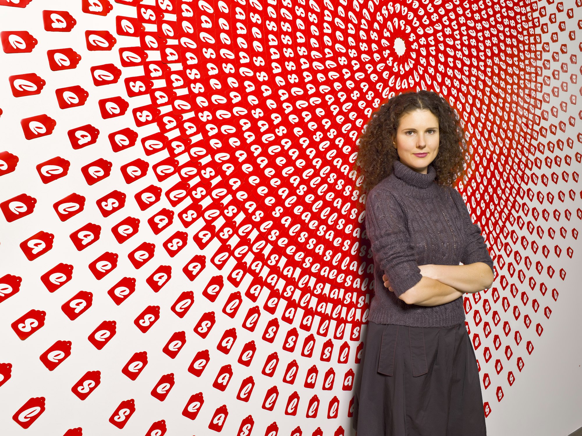 A person with long, centre-parted, curly dark hair, wearing grey polo neck, stands arms crossed against a wall-mounted artwork consisting of many concentric circles of red tags, each with the letter S, A, L or E.