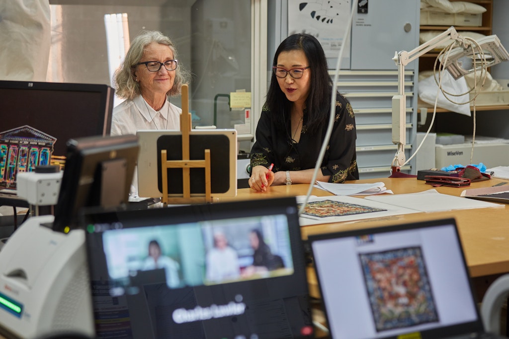 Two people sit at a table looking at a screen. In the foreground are two other screens, one showing video conference attendees and the other a photo of an artwork.