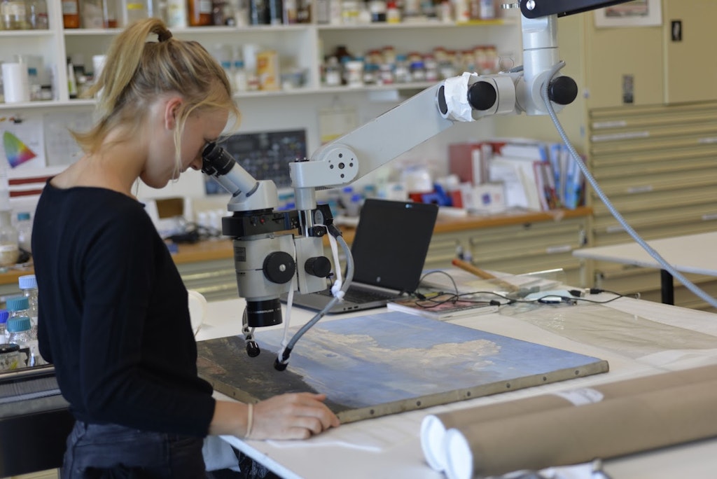 A person stands at a work bench, looking through a large, suspended microscope at a landscape painting, lying flat on the bench.