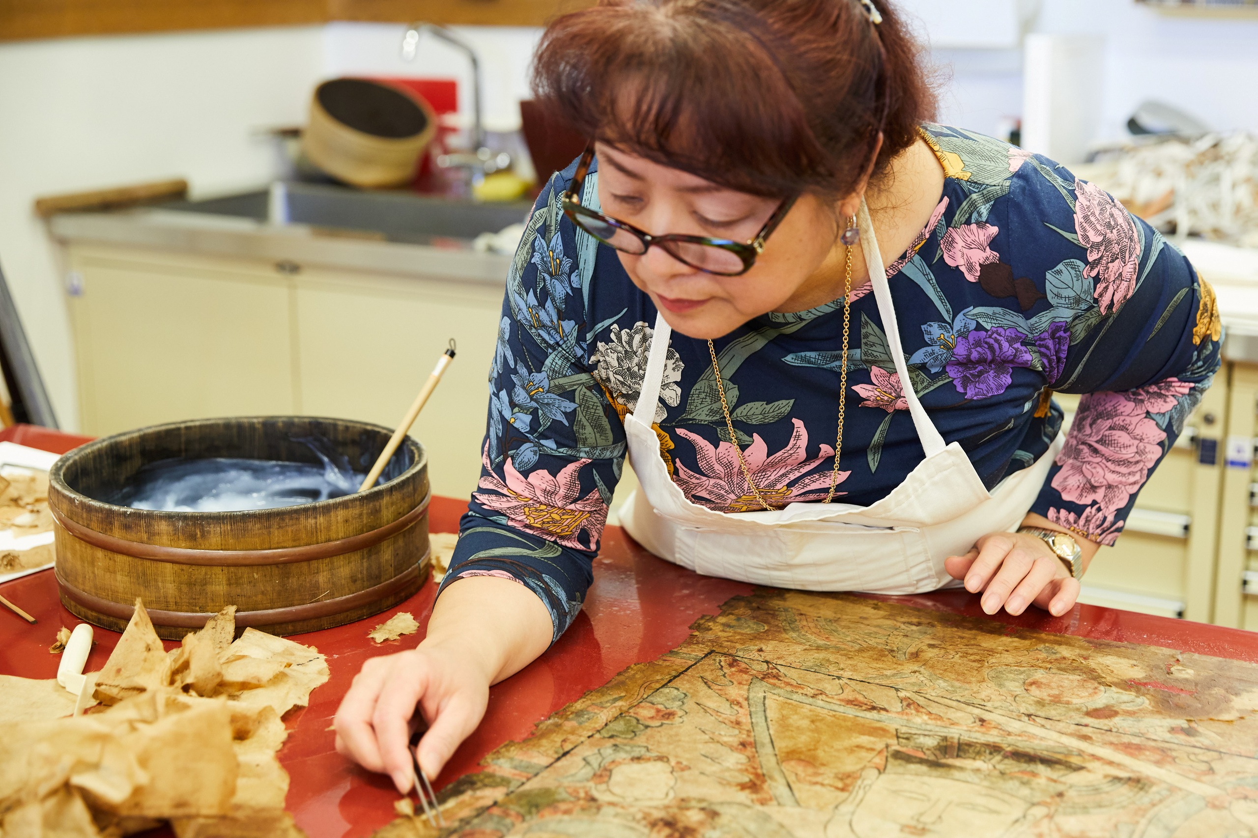 A person leans over a work bench, holding tweezers over a painting lying on the benchtop.