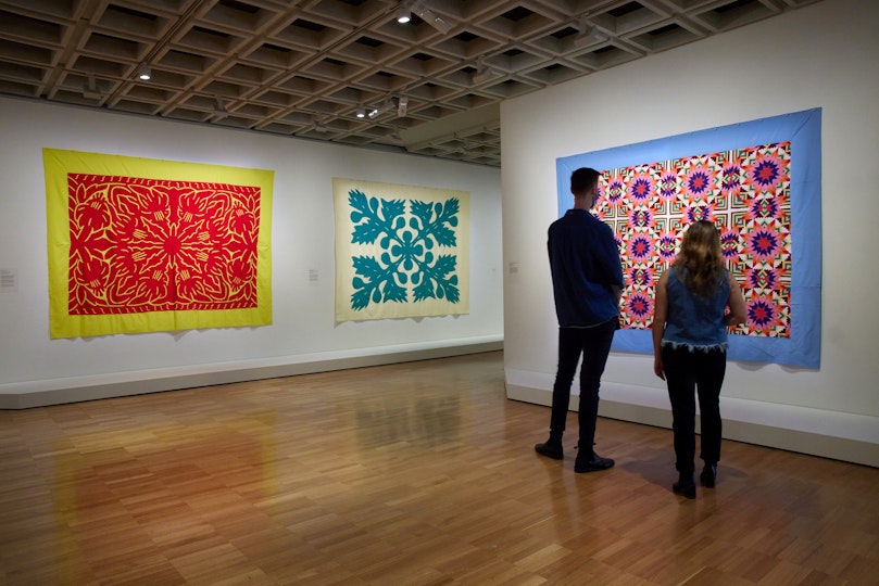 Two people look at a brightly coloured textile artwork on the wall of a gallery, with two other textile works nearby.