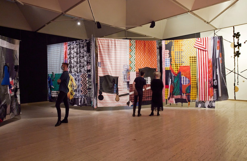People in a gallery look at suspended artworks made from brightly coloured and patterned fabric.