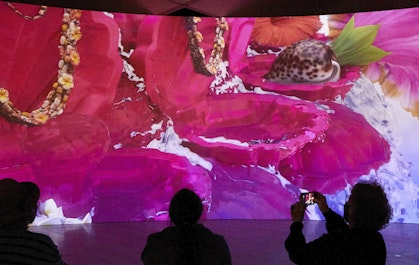 Three people sit in front of a large screen showing a brightly coloured artwork.