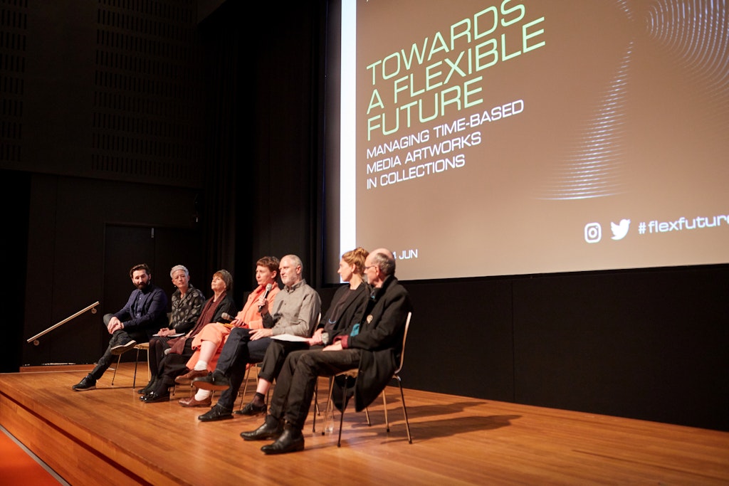 Seven people seated on a stage in front of a screen with text that says 'Towards a flexible future/Managing time-based media artworks in collections'