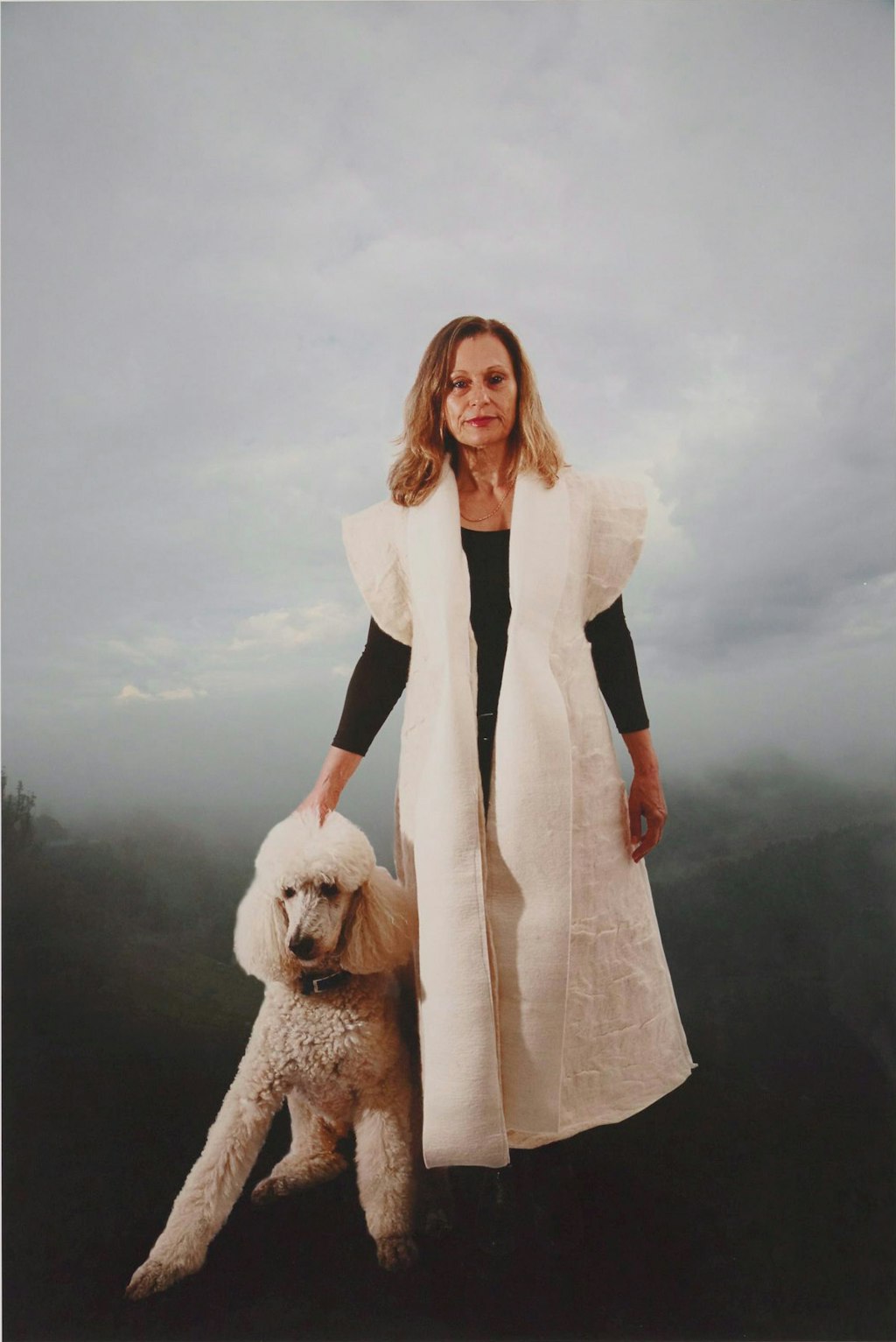 A person wearing a long sleeveless white felt coat stands next to a large white dog.