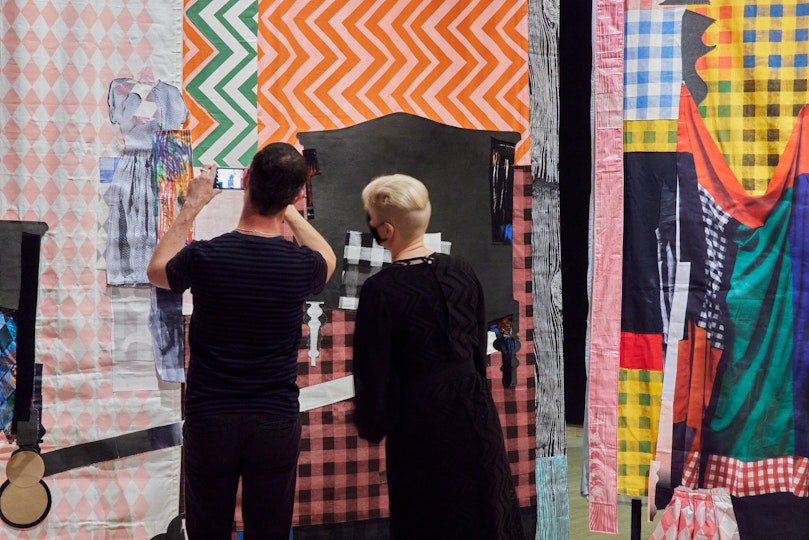 Two people look at an artwork constructed from pieces of coloured, patterned fabric. One of the people is taking a photo with a smartphone.