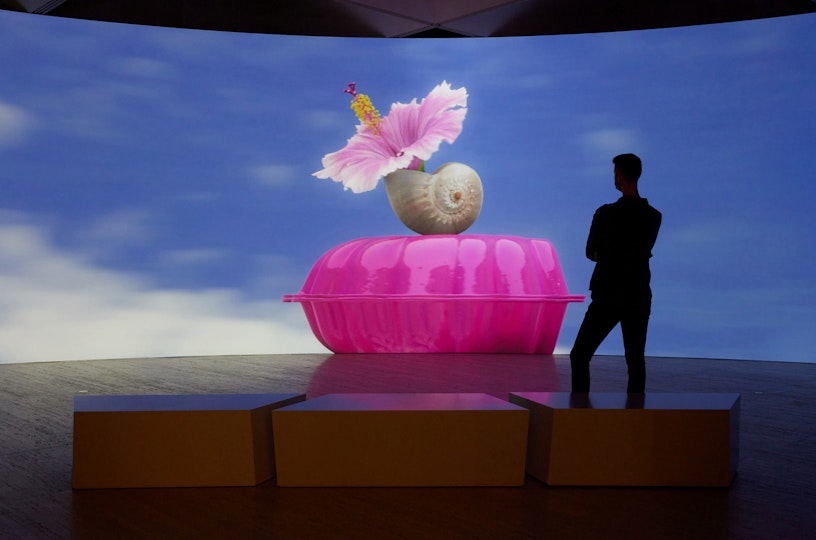 A person stands silhouetted in front of a large curved screen showing, against a blue sky with a few white clouds, a large plastic pink closed clam shell, on which balances a upturned shell holding a flower.
