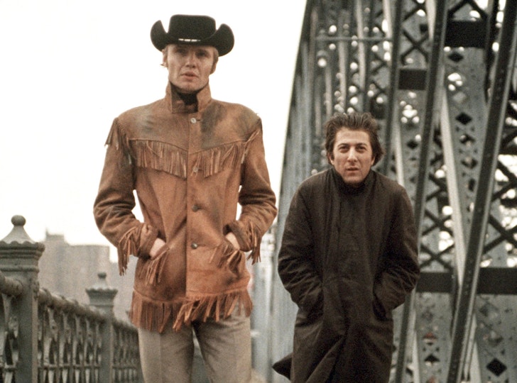 A tall cowboy and a shorter man, both with light skin, in a long coat walk across a bridge.