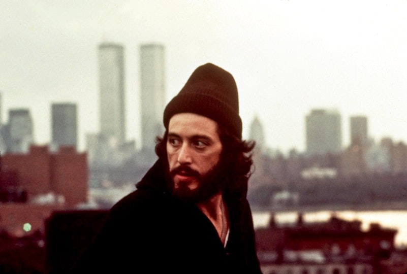 A man with light skin and dark brown hair, moustache and beard is wearing a maroon beanie. Behind him is the New York City skyline.
