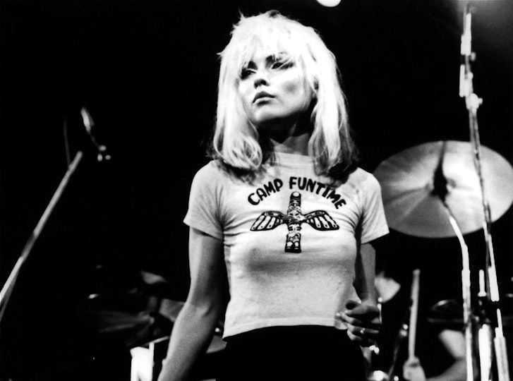 A woman with long bleached hair and light skin stands in front of a drum kit and microphones.